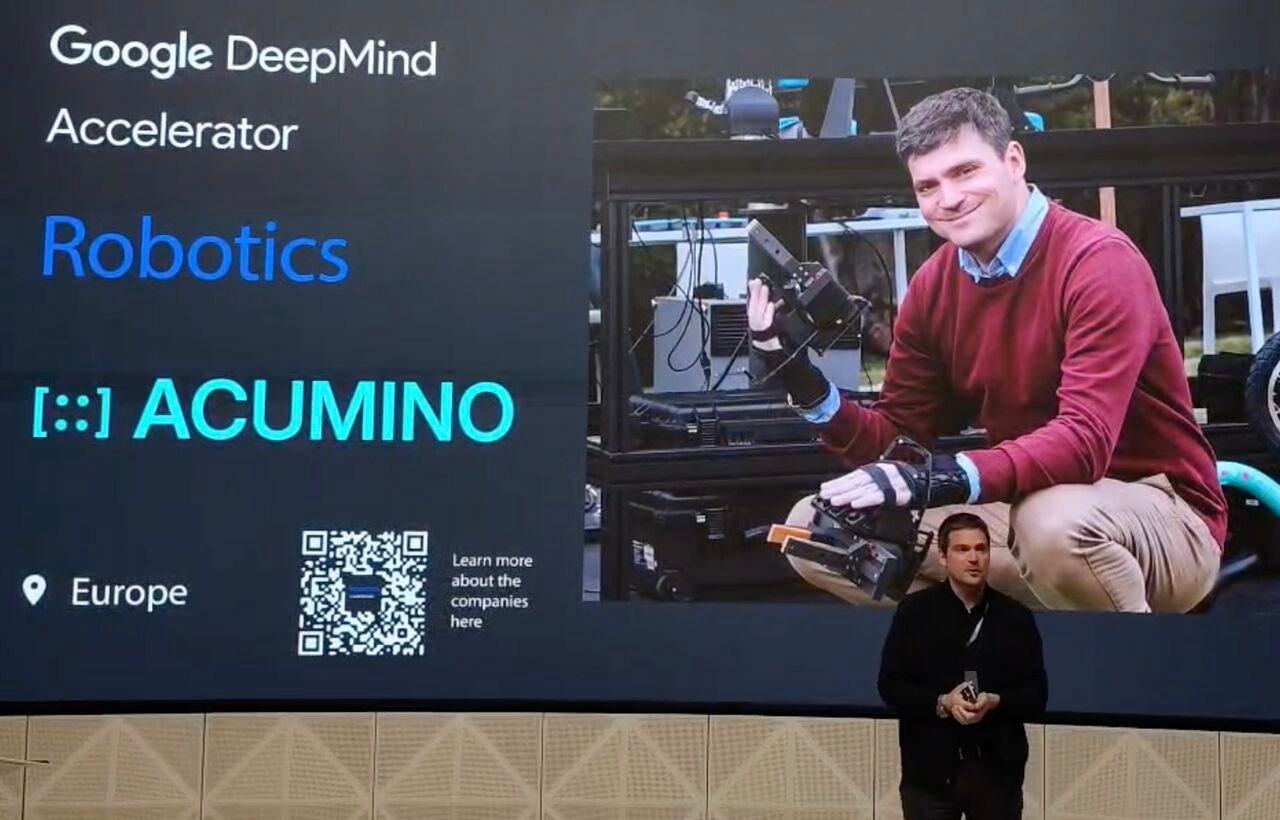 Minas on stage at the Google DeepMind Accelerator Robotics Demo Day in London, with the Acumino slide behind him